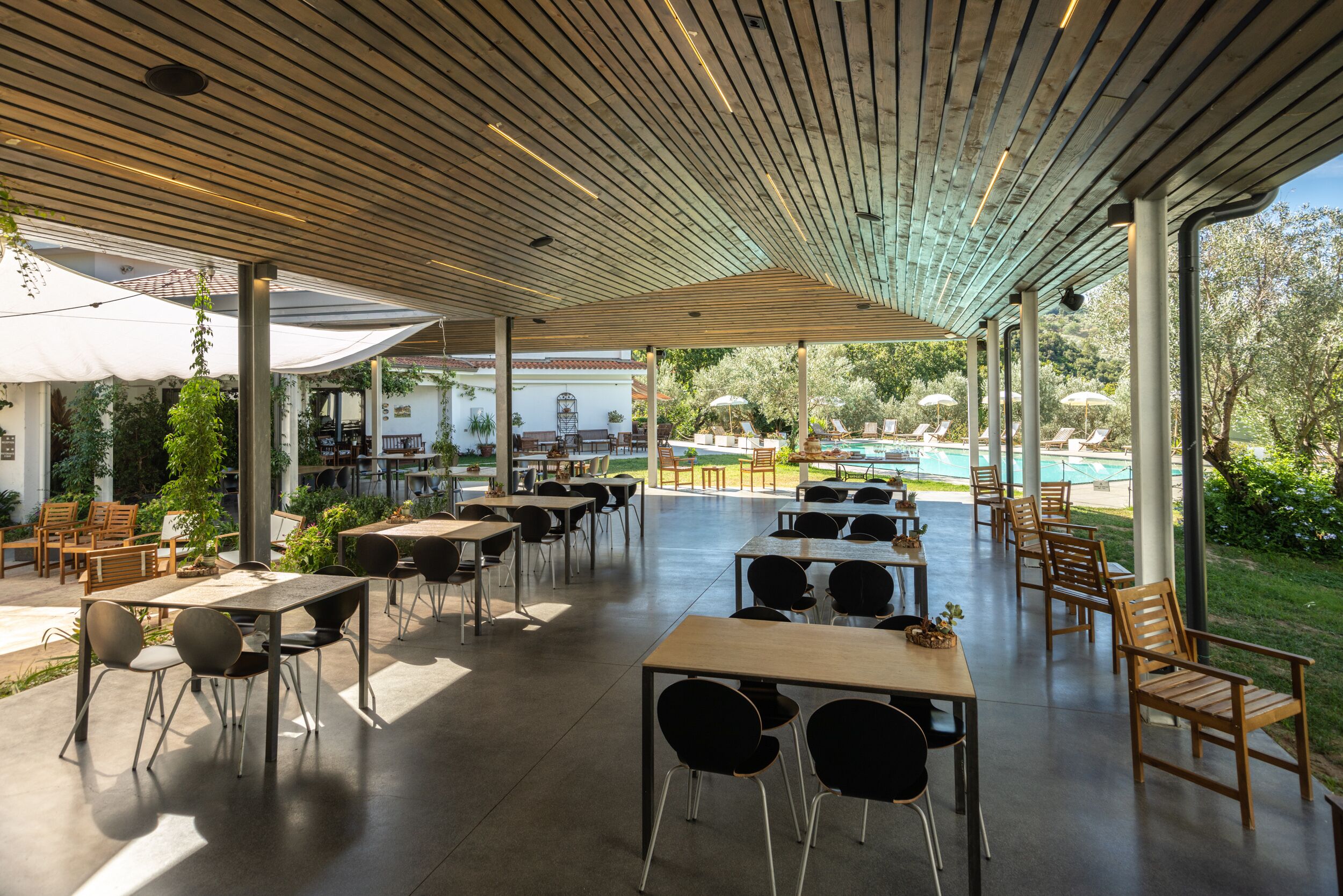 Un patio al aire libre con mesas y sillas, una piscina al fondo y un área de estar techada con sombrillas.