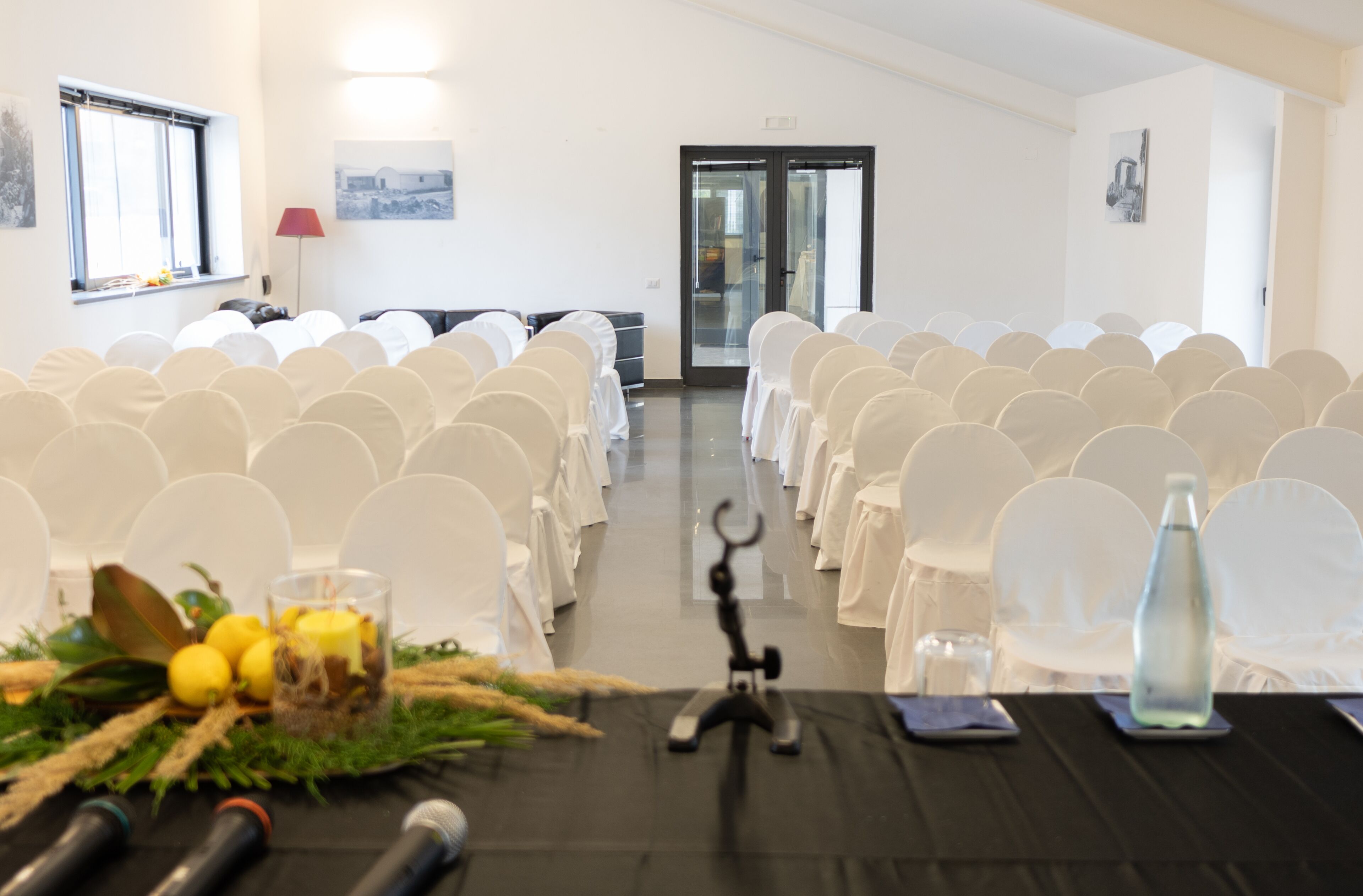 Ein Konferenzraum mit Reihen weißer Stühle, einem schwarzen Tisch mit Mikrofonen und einer Glasflasche auf dem Tisch.