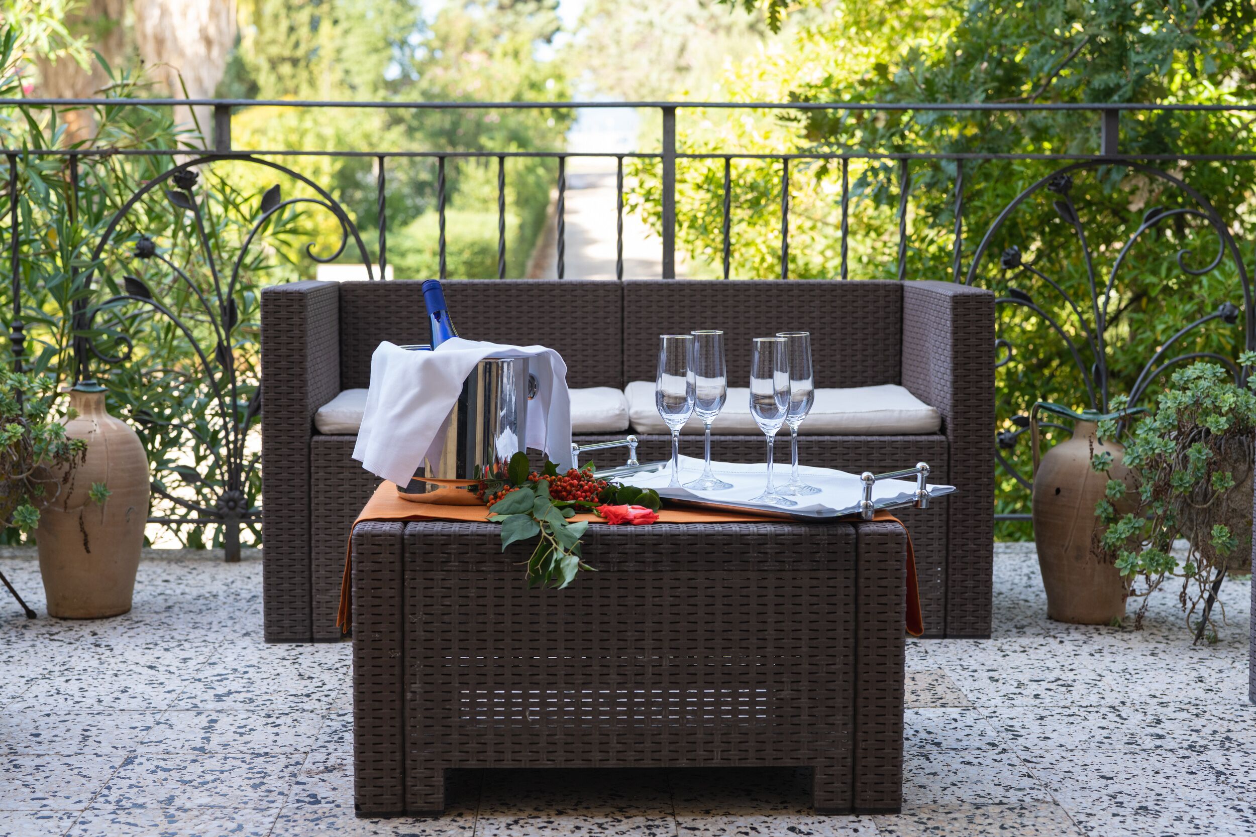 Eine Korbsitzgruppe mit Kissen, ein Tisch mit Weingläsern und einer Karaffe, auf einer Terrasse mit Geländer und Grünpflanzen im Hintergrund.