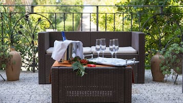Eine Korbsitzgruppe mit Kissen, ein Tisch mit Weingläsern und einer Karaffe, auf einer Terrasse mit Geländer und Grünpflanzen im Hintergrund.