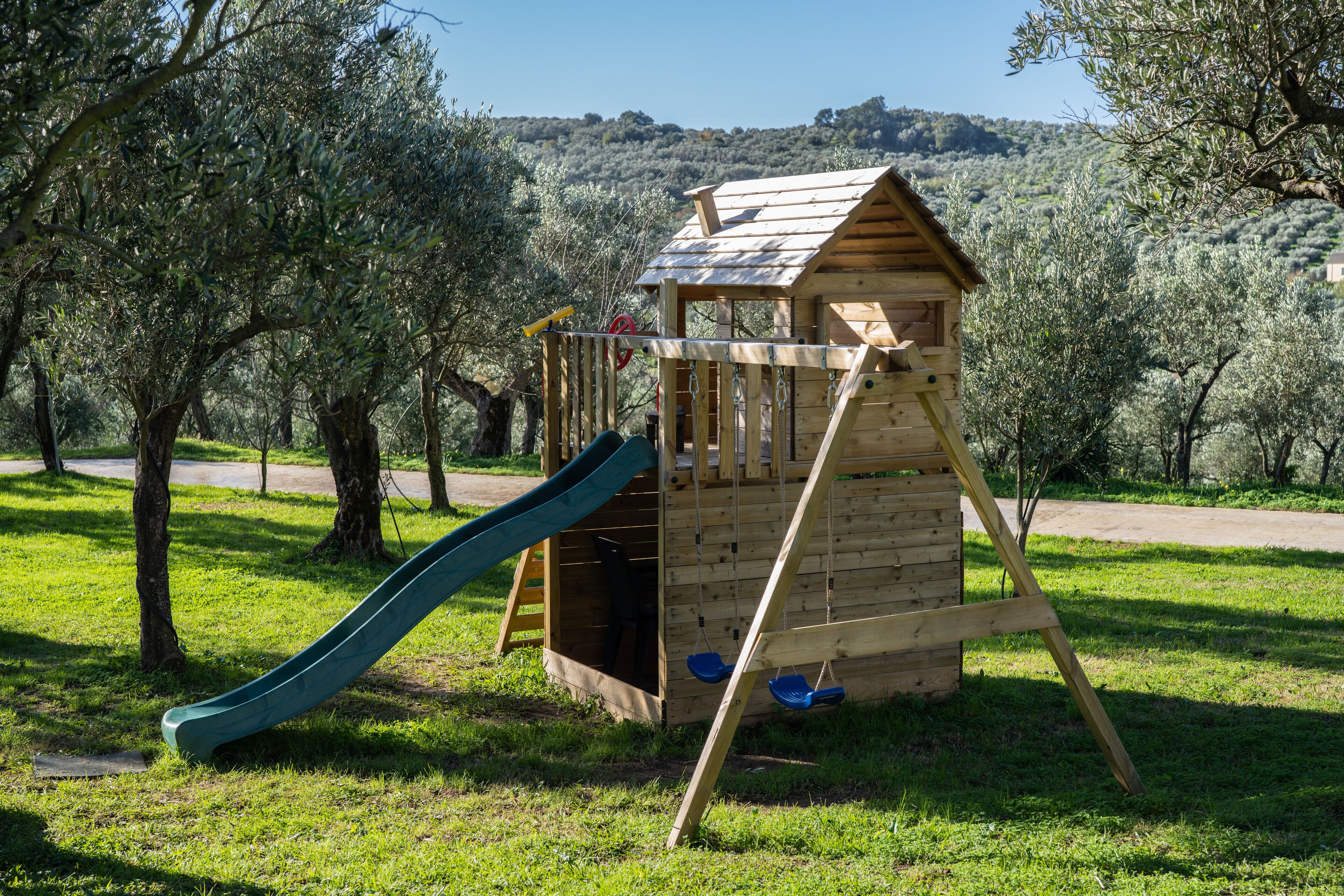 A wooden playhouse with a slide in a grassy park area with olive trees.