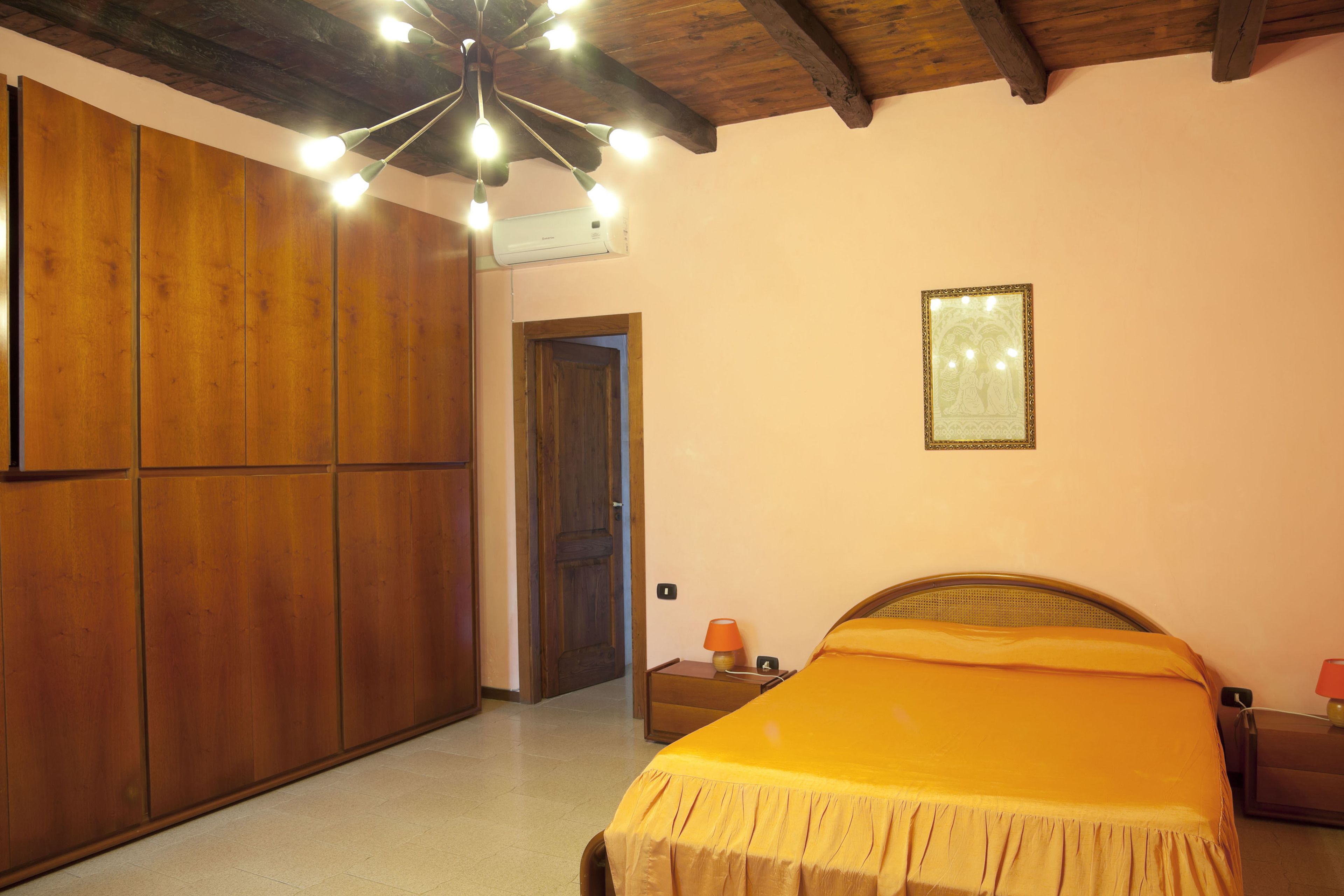 A bedroom with a wooden wardrobe, a bed with a yellow bedspread, a nightstand with a lamp, and a ceiling light fixture.