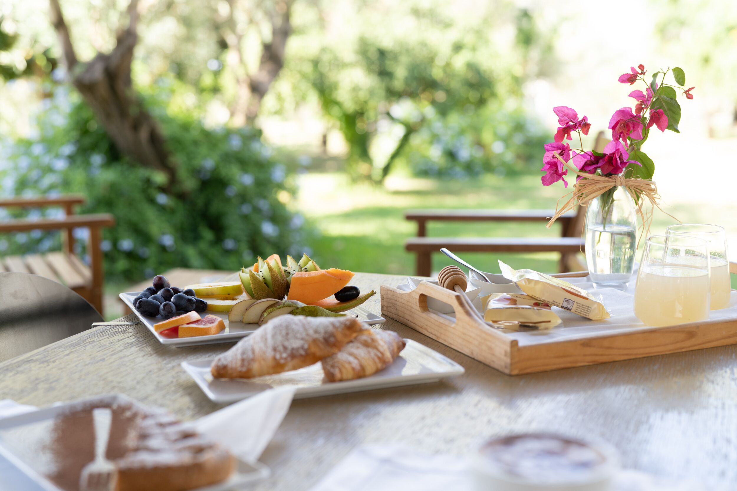 A picnic setup with food and drinks on a wooden table, surrounded by greenery.