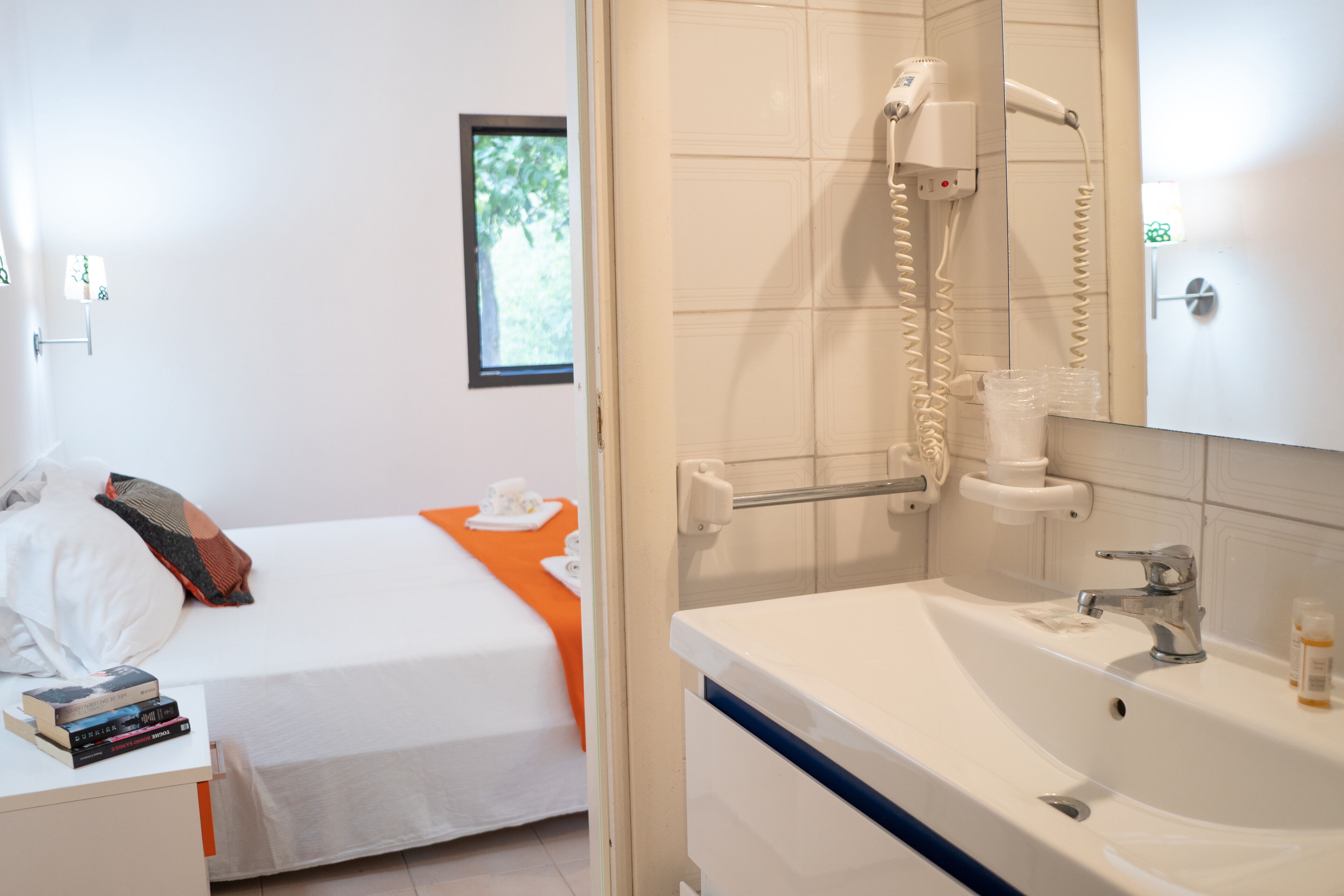 Habitación de hotel con cama, mesita de noche con libros, ventana con vistas, baño con lavabo y espejo, y ducha con secador.