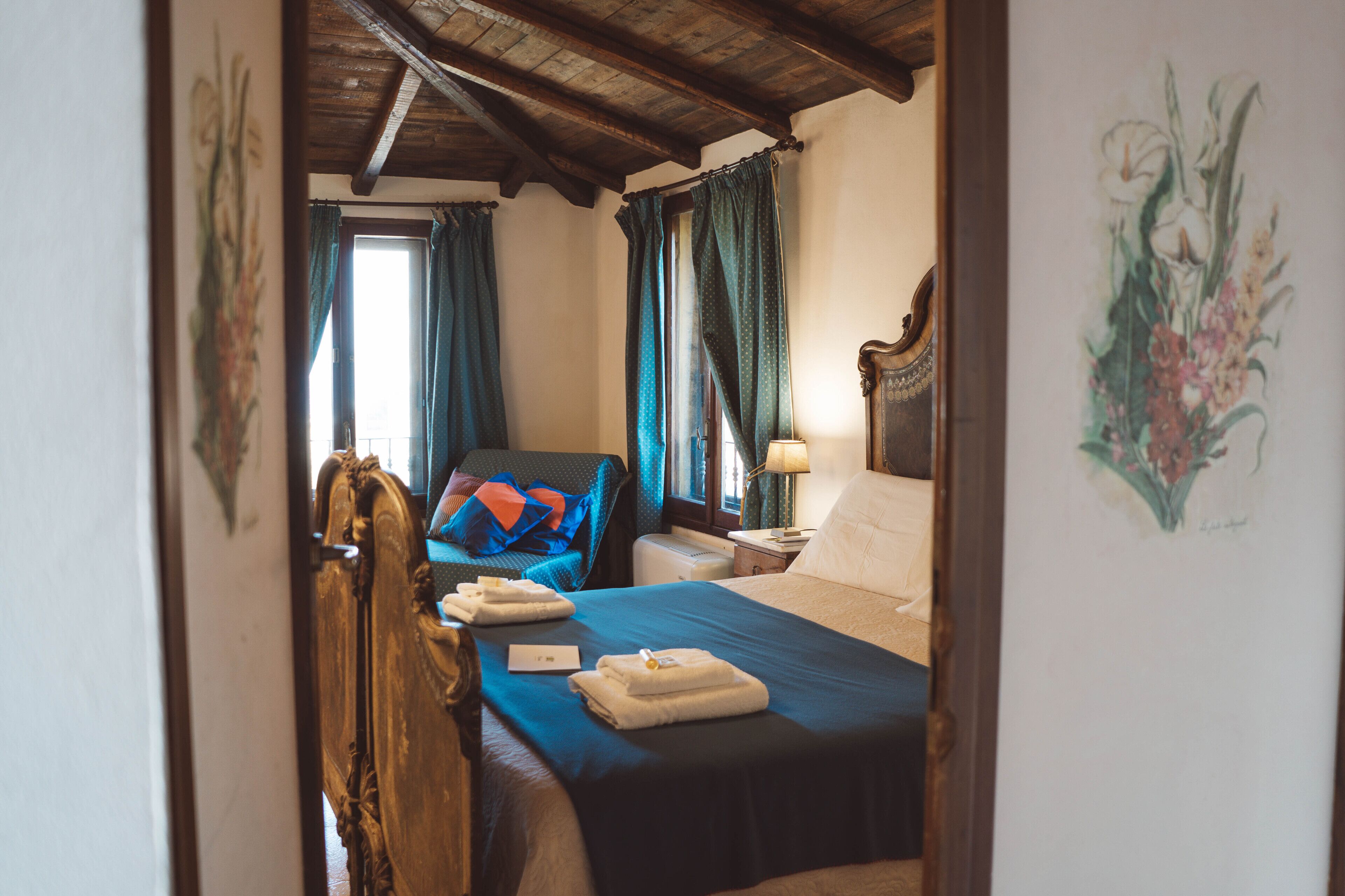 Una habitación de hotel con techo de madera, una cama con cubrecama azul, una silla con cojín azul, una mesita de noche con lámpara y un cuadro en la pared.