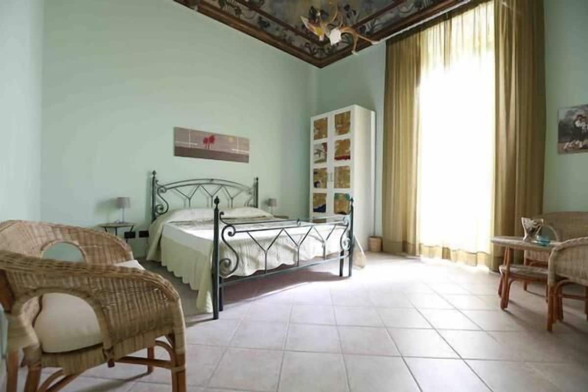 A bedroom with a bed, two wicker chairs, a wardrobe, and a window with curtains.
