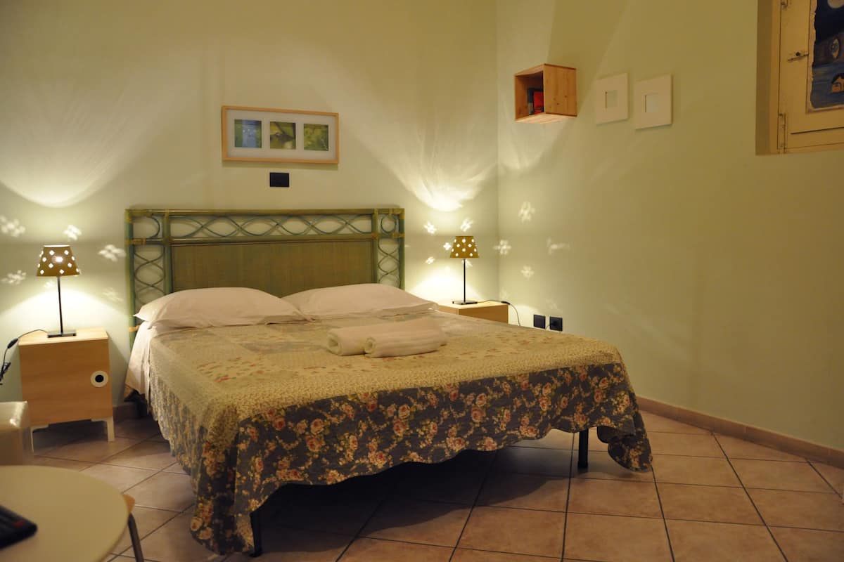 A neatly made bed with a floral bedspread and pillows, two bedside tables with lamps, a small wooden shelf, and a framed picture on the wall.