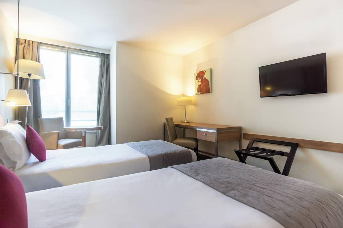 Une chambre d’hôtel avec deux lits, un bureau, une chaise, une télévision et une fenêtre avec des rideaux.