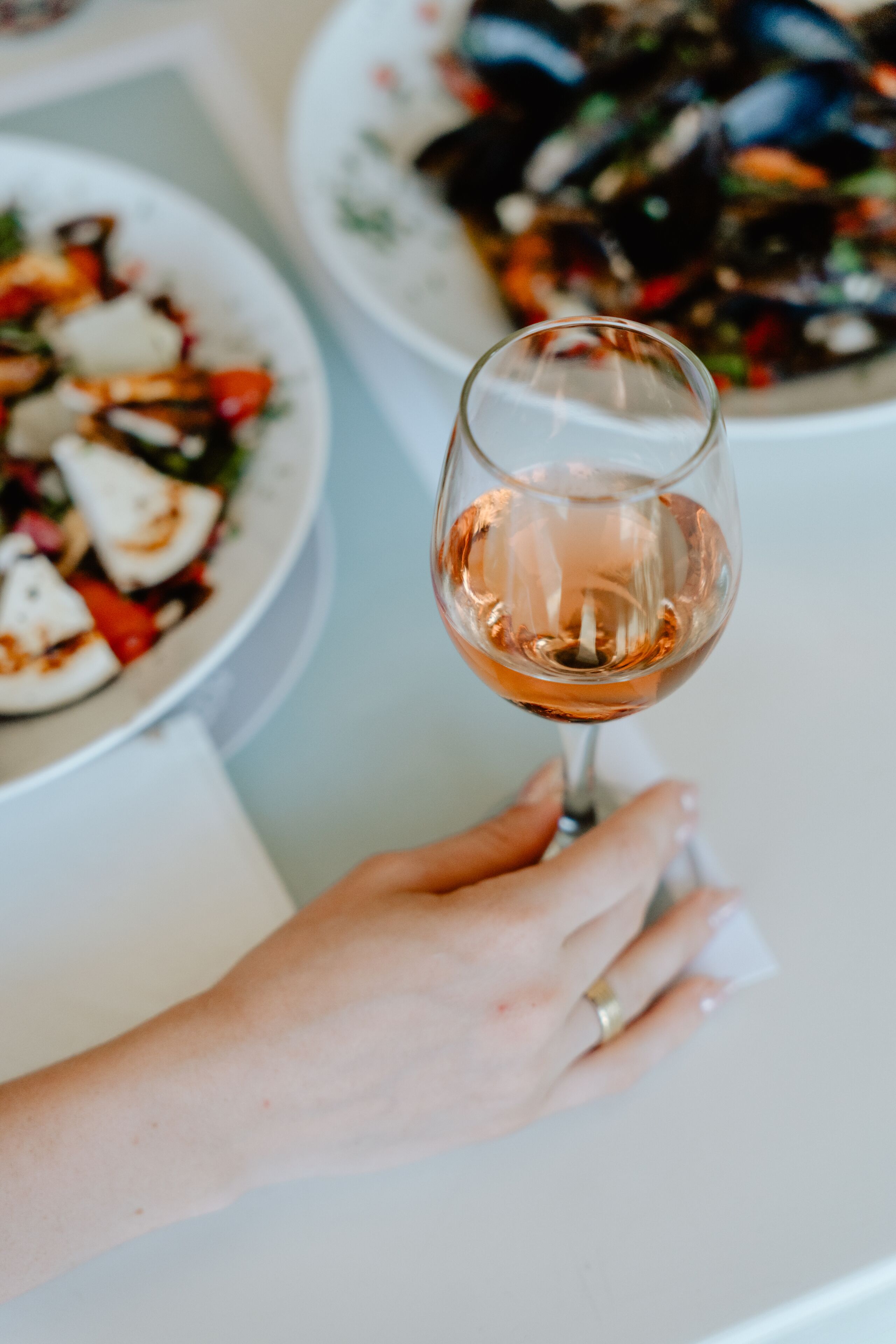 A hand holding a wine glass with a rose-colored liquid, a plate of food with vegetables and cheese, and another plate with a seafood dish.