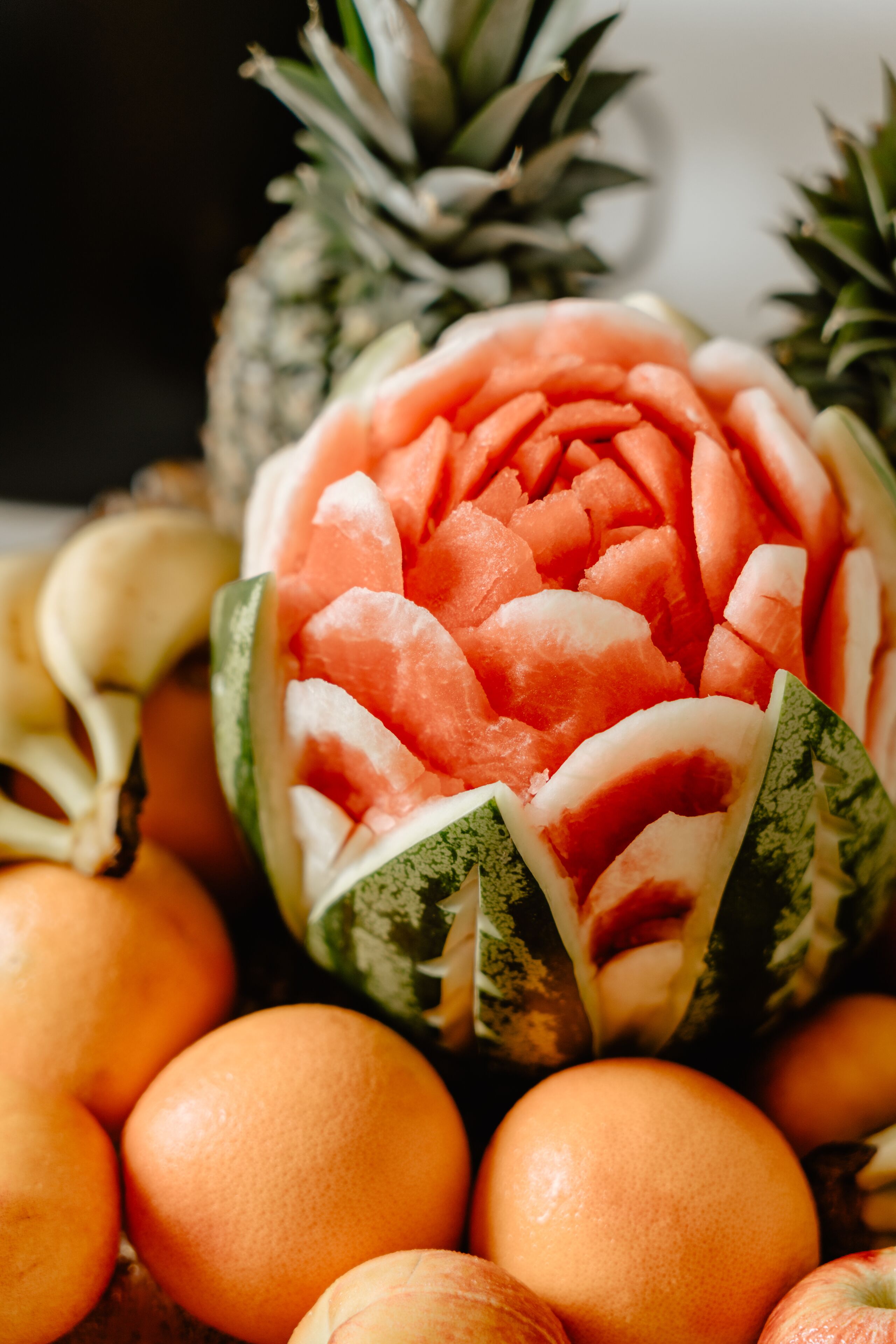 A watermelon carved into a floral design surrounded by oranges and a pineapple.