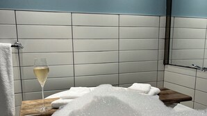 A bathtub filled with bubbles, a glass of champagne, and a wooden shelf with towels.
