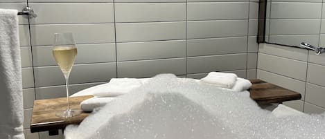 A bathtub filled with bubbles, a glass of champagne, and a wooden shelf with towels.