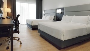 A hotel room with two beds, a desk with a lamp, a chair, and a large window with curtains.