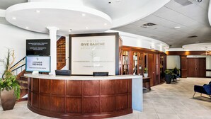 A hotel reception area with a wooden reception desk, a large sign for Hotel Rive Gauche, and a staircase in the background.