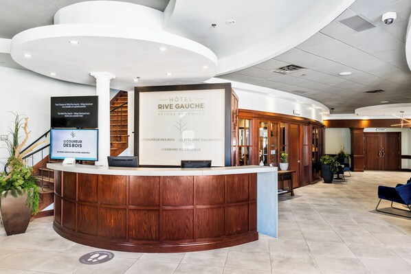 A hotel reception area with a wooden reception desk, a large sign for Hotel Rive Gauche, and a staircase in the background. - Hôtel Rive Gauche (Beloeil)