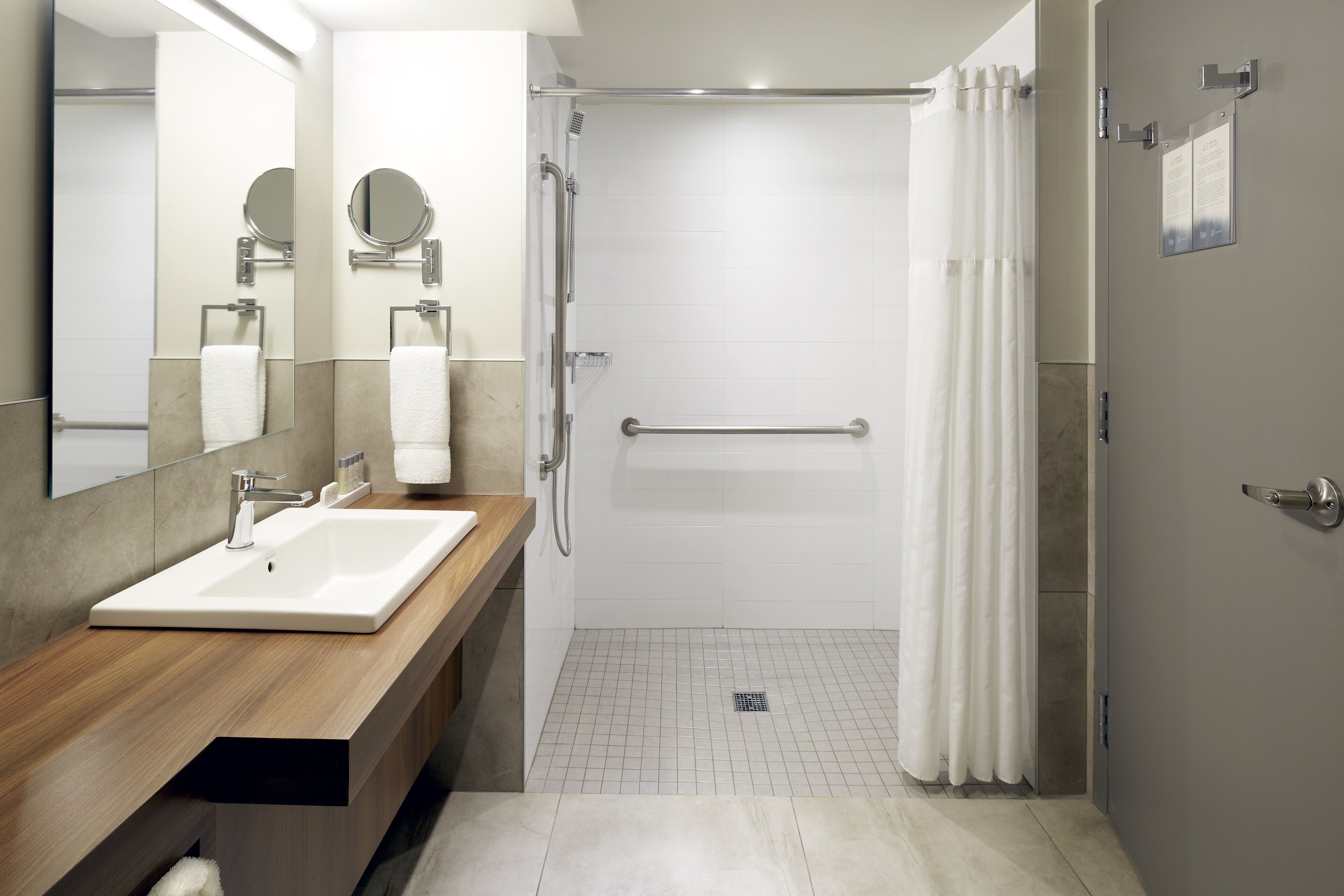 A modern bathroom with a wooden vanity, a large mirror, a shower with a glass door, and a tiled floor.