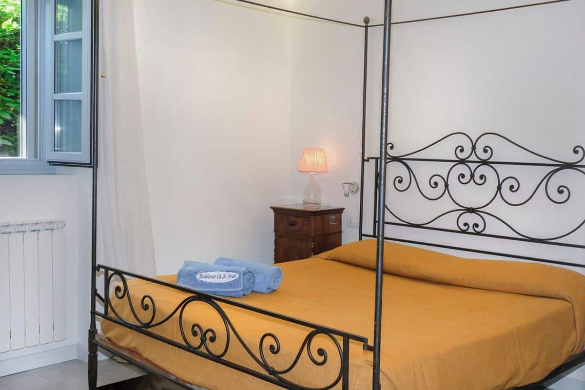 A four-poster bed with a yellow bedspread and blue towels, a bedside table with a lamp, and a view of greenery outside.