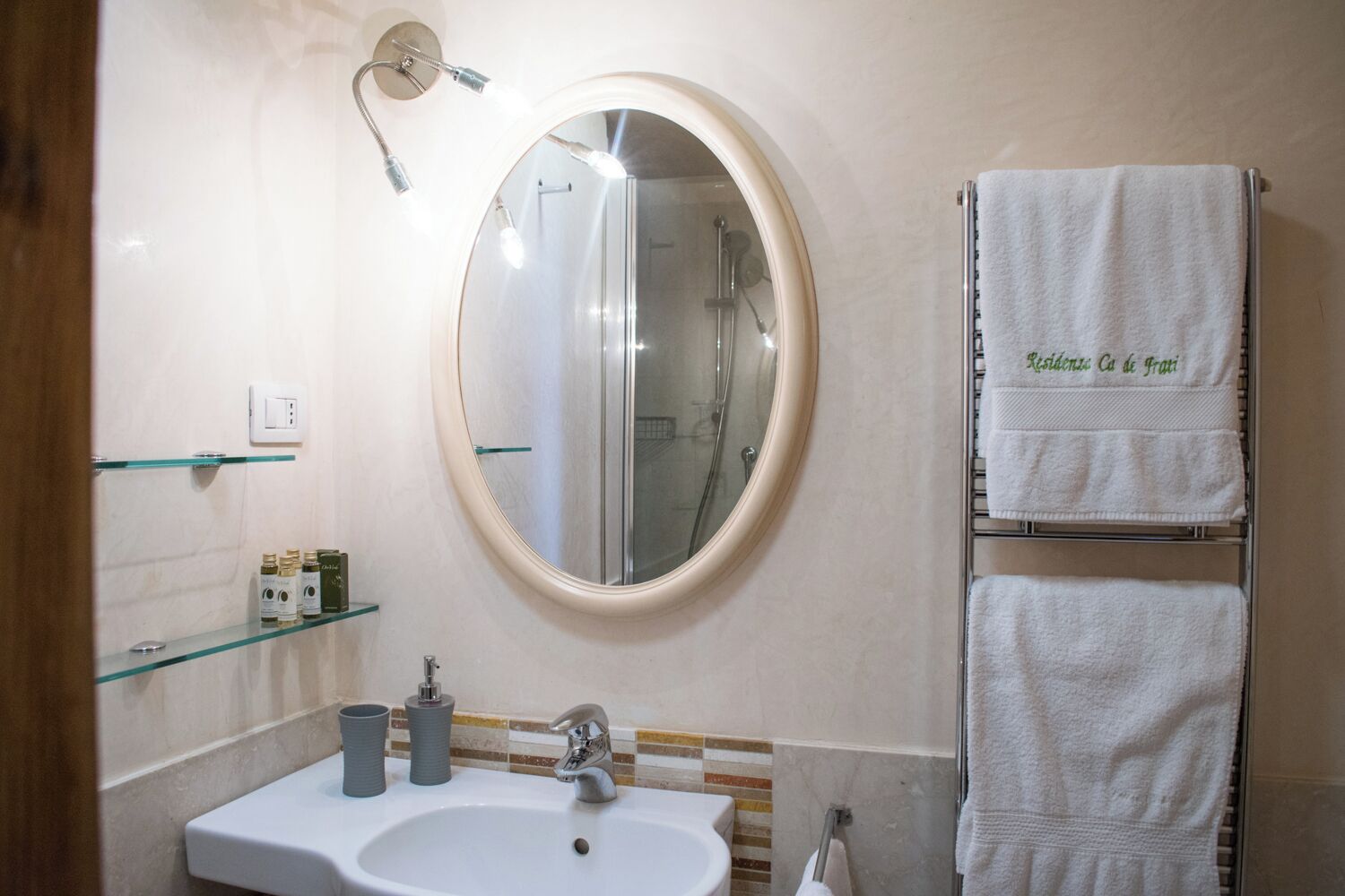 Een badkamer met een ronde spiegel, een wastafel, een glazen plankje met toiletartikelen en een handdoekrek met twee handdoeken.
