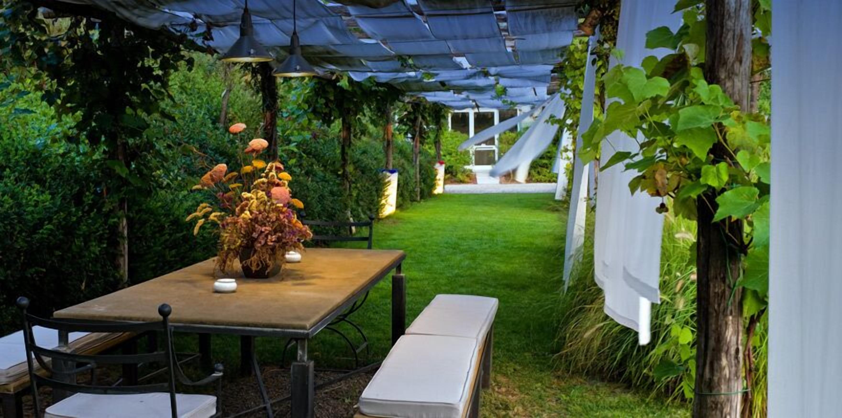 Un patio al aire libre con una mesa y bancos, rodeado de follaje y una zona cubierta con luces colgantes.