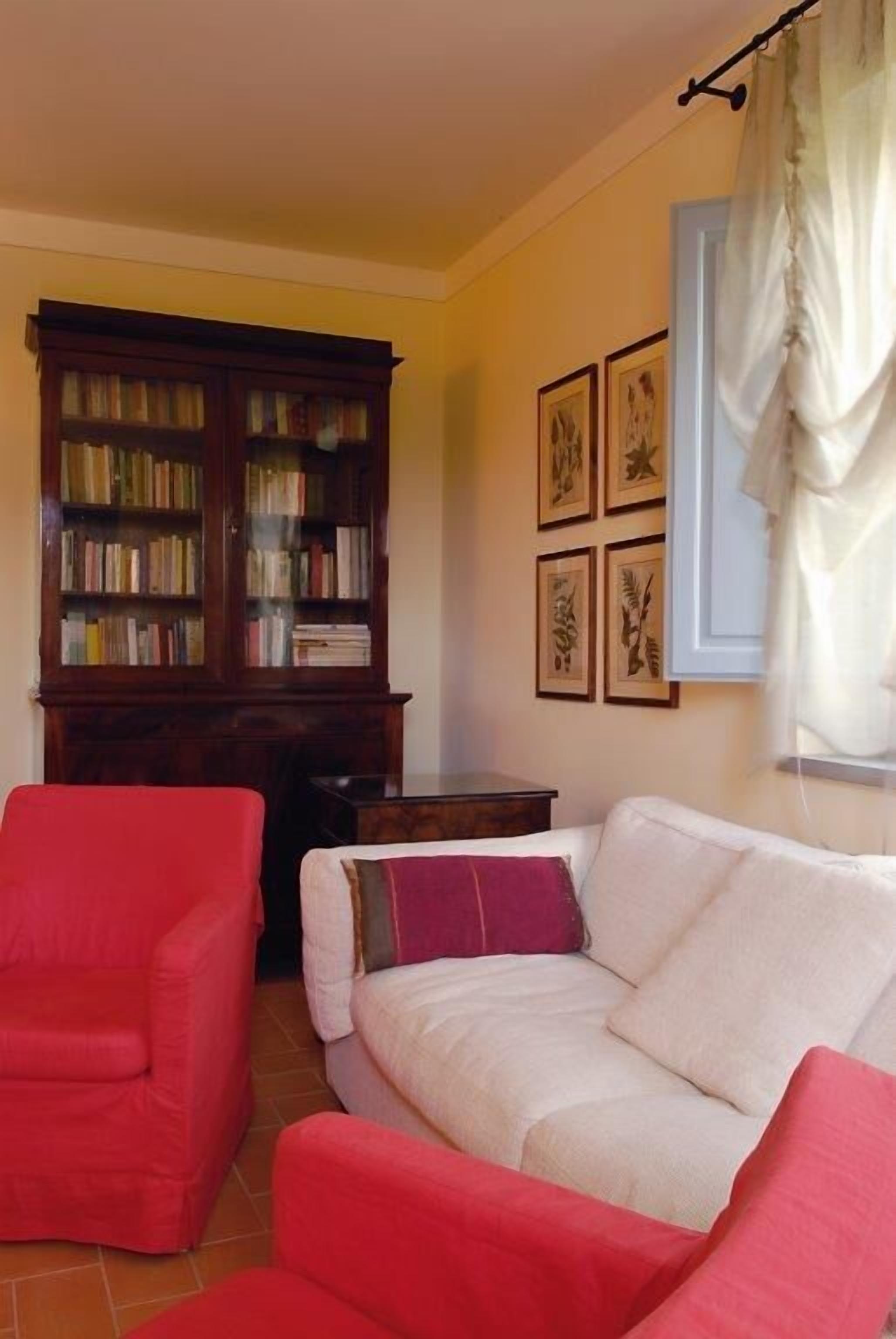 A living room with a sofa, a red armchair, a wooden bookshelf, and framed botanical prints on the wall.