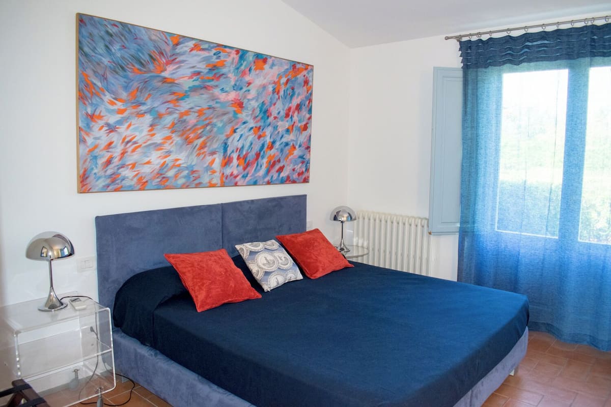 A bedroom with a large bed, a bedside table, a lamp, a radiator, and a window with curtains.