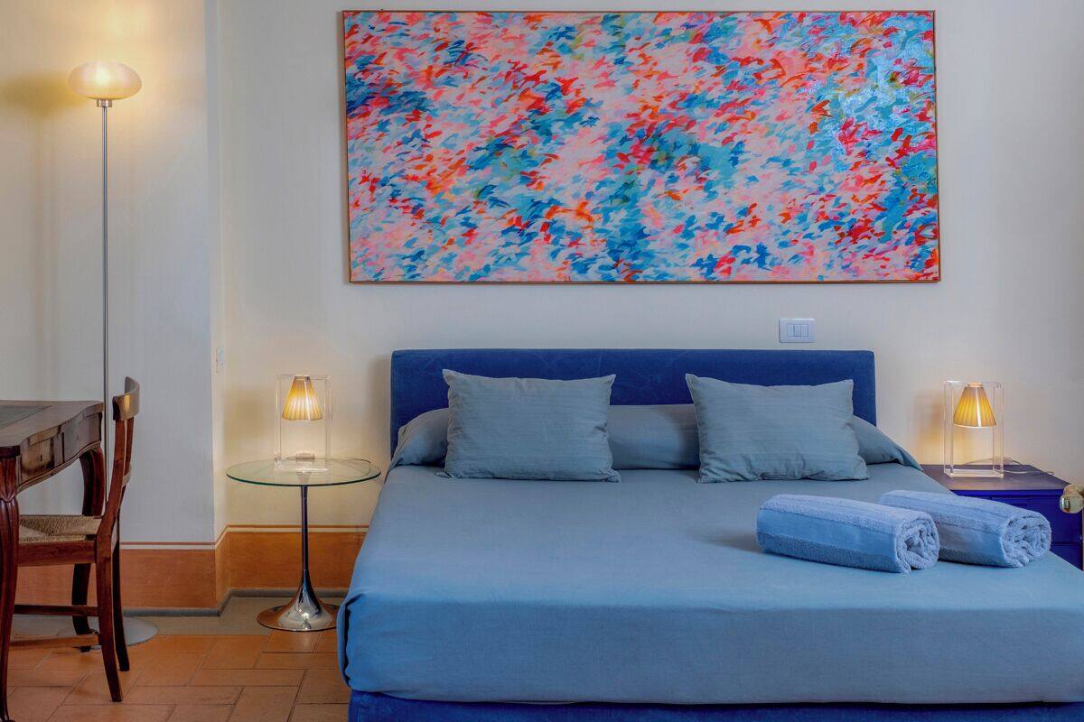 A bedroom with a bed, pillows, a blue blanket, a glass table with a lamp, a chair, and a colorful abstract painting on the wall.