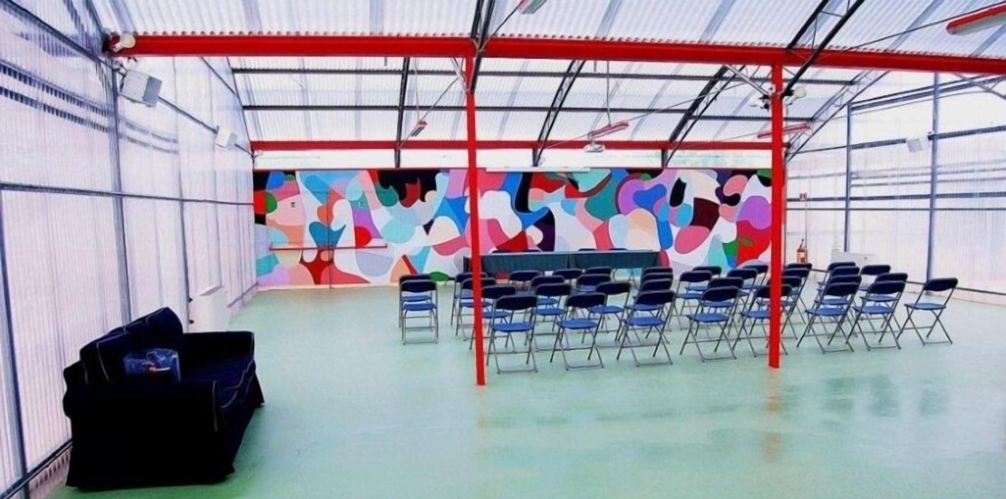 Een ruimte binnenshuis met een rode metalen constructie, rijen blauwe opklapbare stoelen en een kleurrijke abstracte muurschildering op de achterwand.