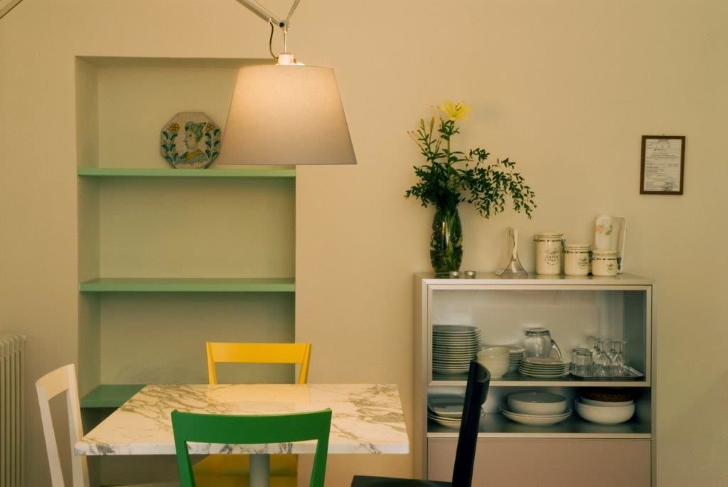 A dining area with a table, chairs, a hanging lamp, a vase with flowers, and shelves with dishes and jars.