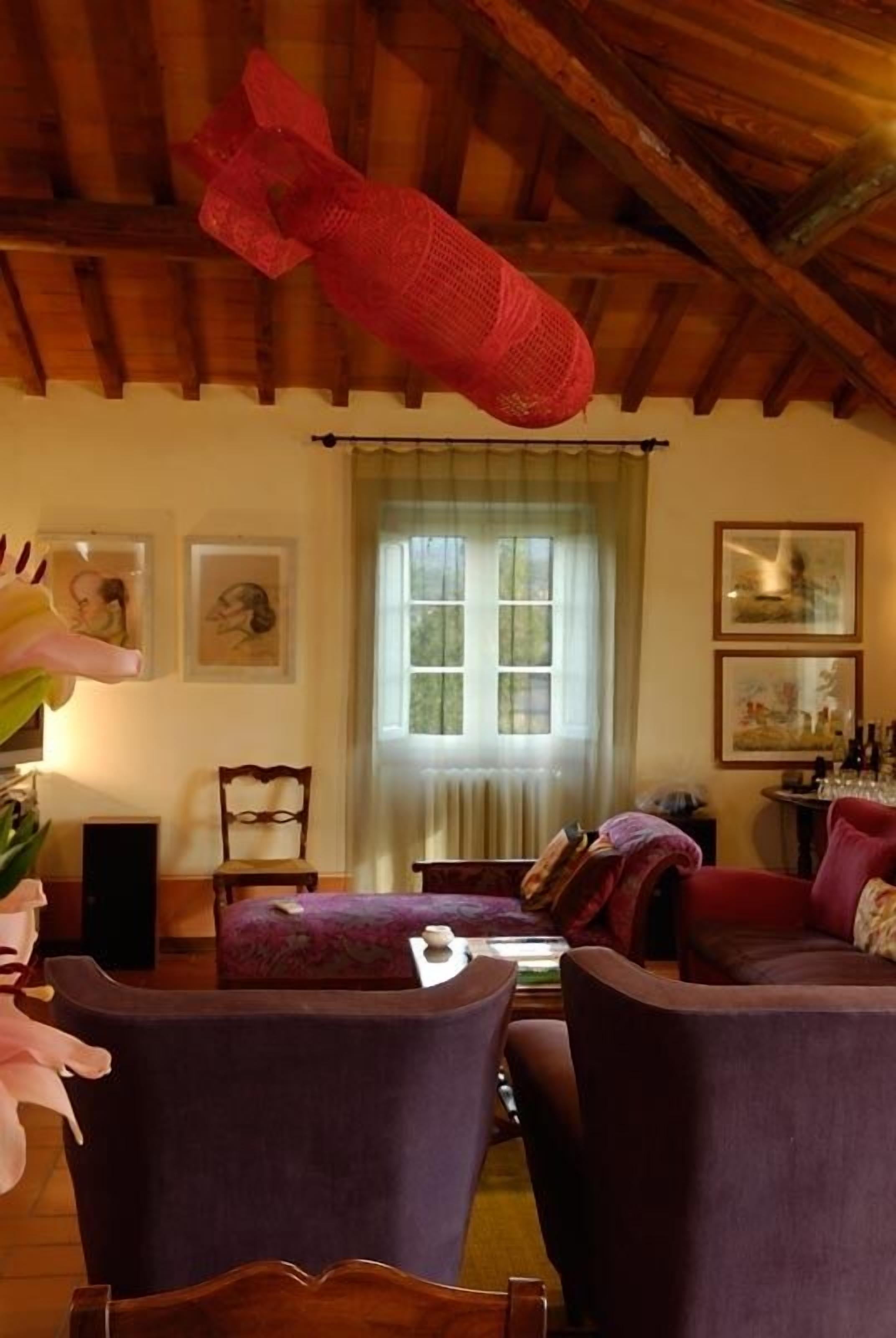 A cozy living room with a high wooden ceiling, a window with curtains, and a fireplace. There are two purple armchairs and a wooden table with a vase of flowers.