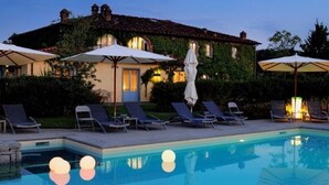 Uma área de piscina com espreguiçadeiras e guarda-sóis, piscina e um edifício com telhado telhado.