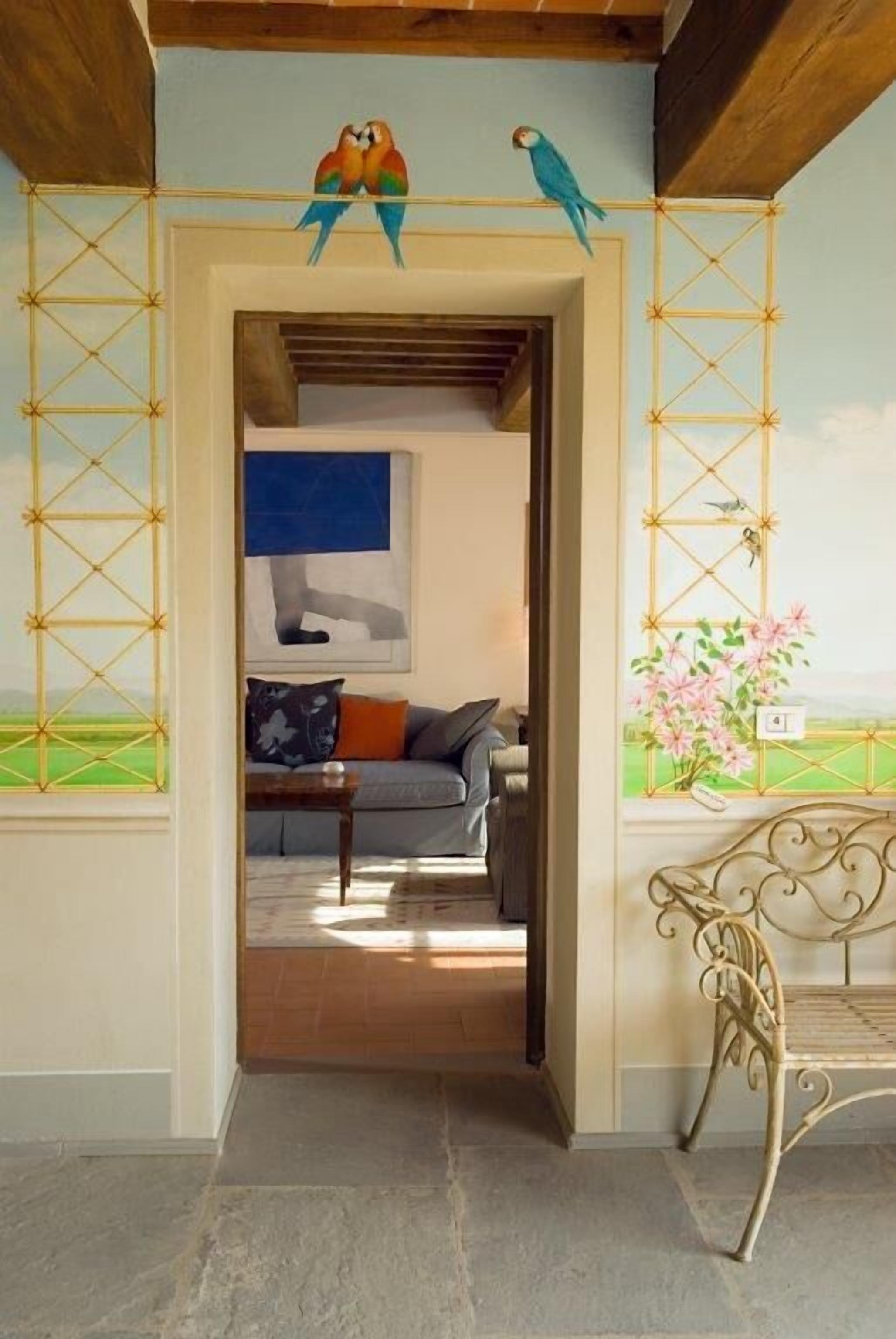 Une pièce avec un plafond en bois, un canapé avec des coussins, un banc en métal et un mur orné d’une fenêtre et d’un tableau représentant des oiseaux.