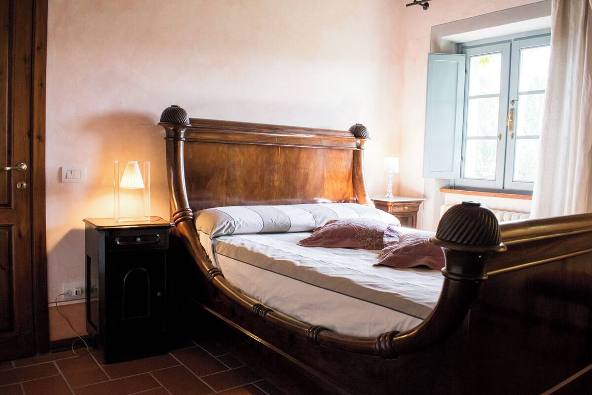 A wooden four-poster bed with a nightstand and lamp, a window with curtains, and a door.