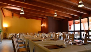 A restaurant with multiple tables set for dining, wooden beams on the ceiling, and large windows with curtains.