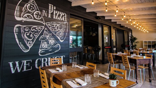 Interior de restaurante con vigas de madera en el techo, luces colgantes y un mural en la pared con porciones de pizza. Mesas dispuestas con sillas y un menú a la vista.