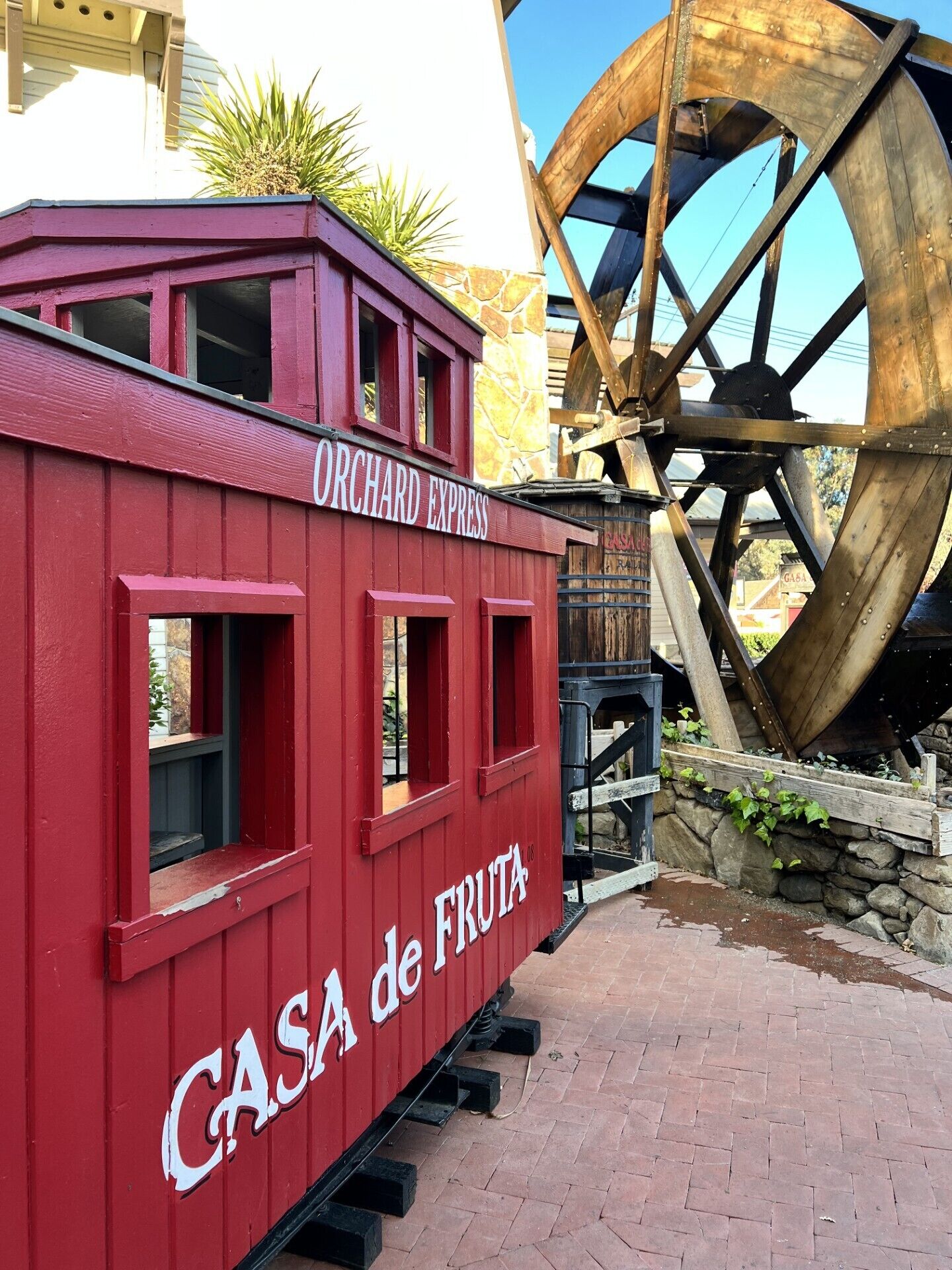 A red train car with "Orchard Express" and "Casa de Fruta" written on it, parked near a large wooden water wheel.