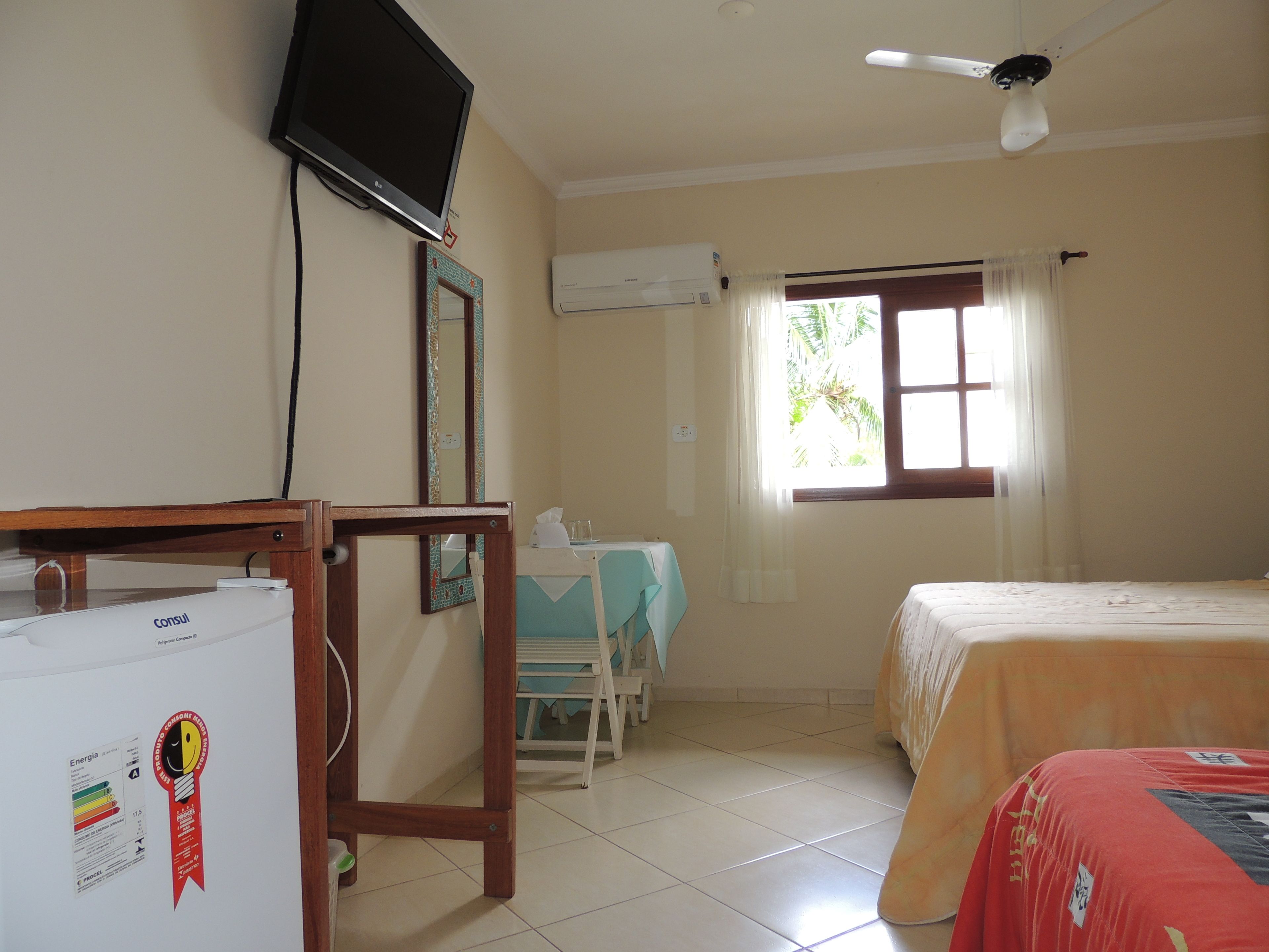 A hotel room with a bed, a desk with a chair, a television, a ceiling fan, and an air conditioning unit.