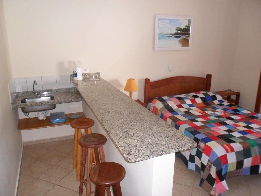 A compact room with a kitchenette, a bed with a colorful quilt, and a small table with a lamp.
