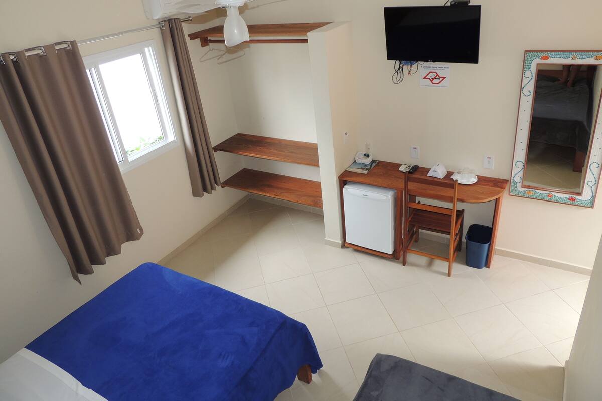 A compact hotel room with a bed, a desk with a computer, a small fridge, a TV, and a mirror.