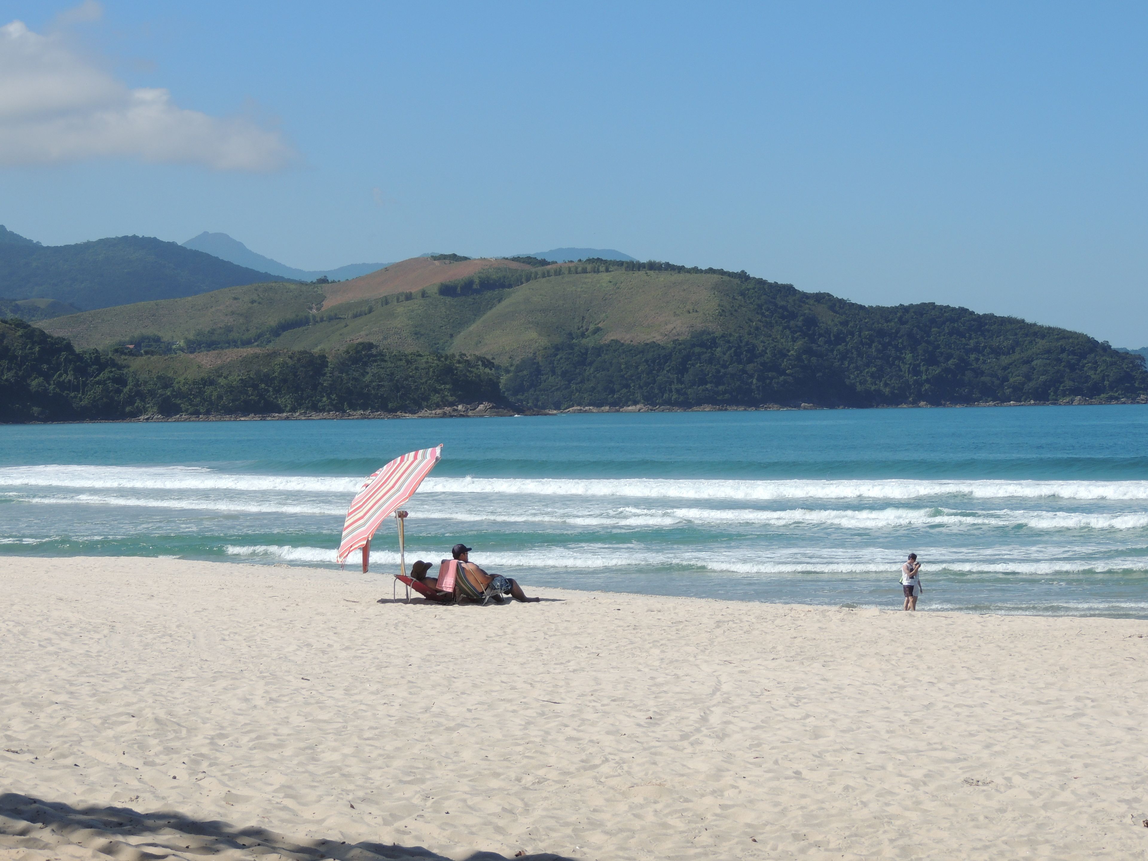 A beach with a striped umbrella, a person lying on a beach chair, and another person standing in the water.