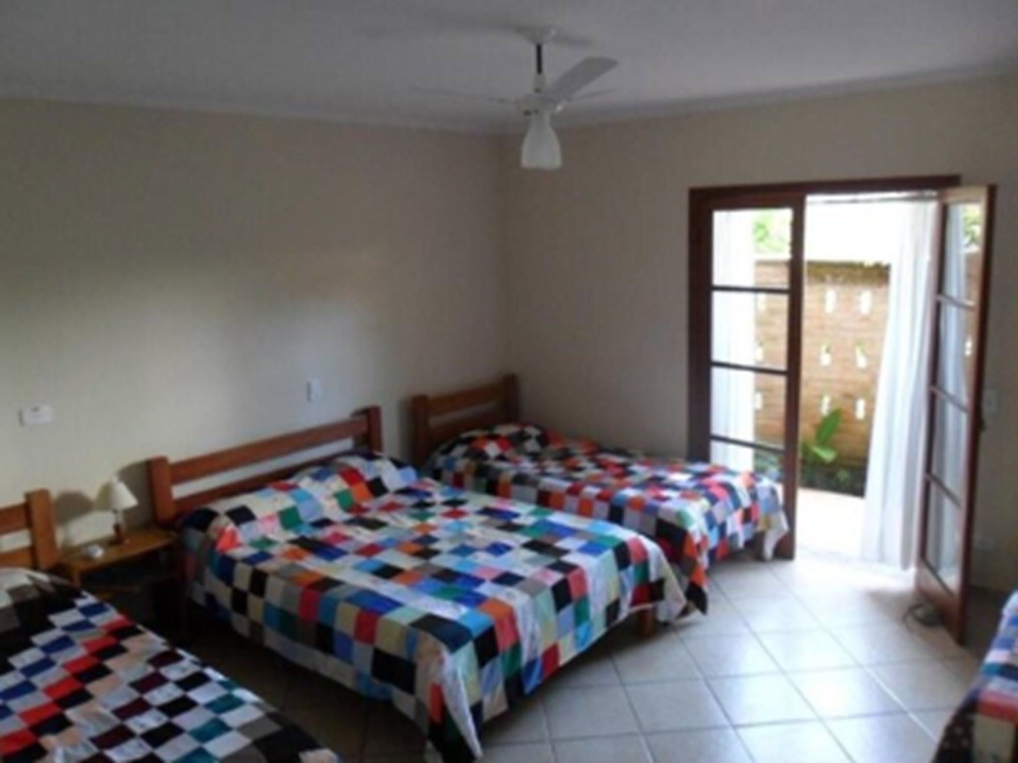 A room with three beds, each with a colorful quilt, a ceiling fan, and a window with a view of greenery outside.