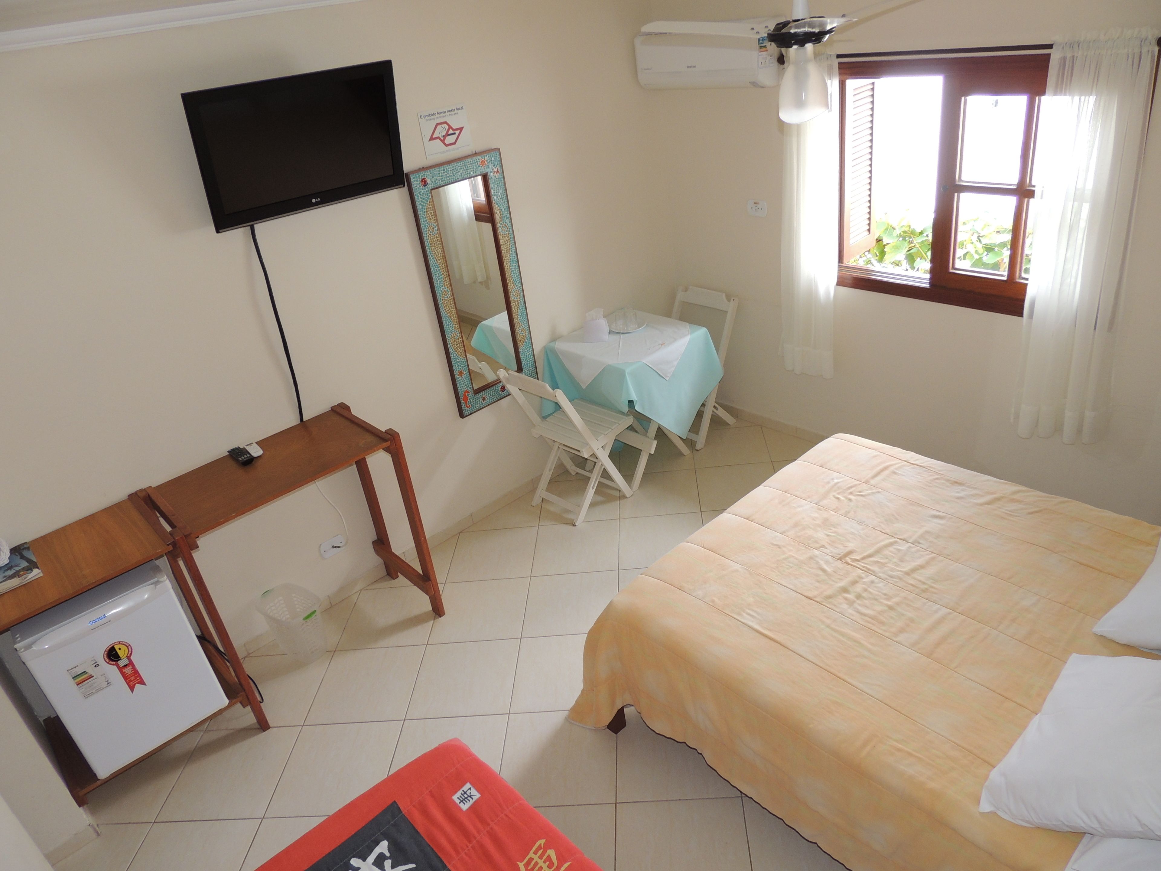 A bedroom with a bed, a desk with a refrigerator, a chair, a table with a tablecloth, a TV mounted on the wall, and an air conditioning unit.
