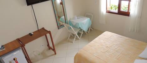 A bedroom with a bed, a desk with a refrigerator, a chair, a table with a tablecloth, a TV mounted on the wall, and an air conditioning unit.
