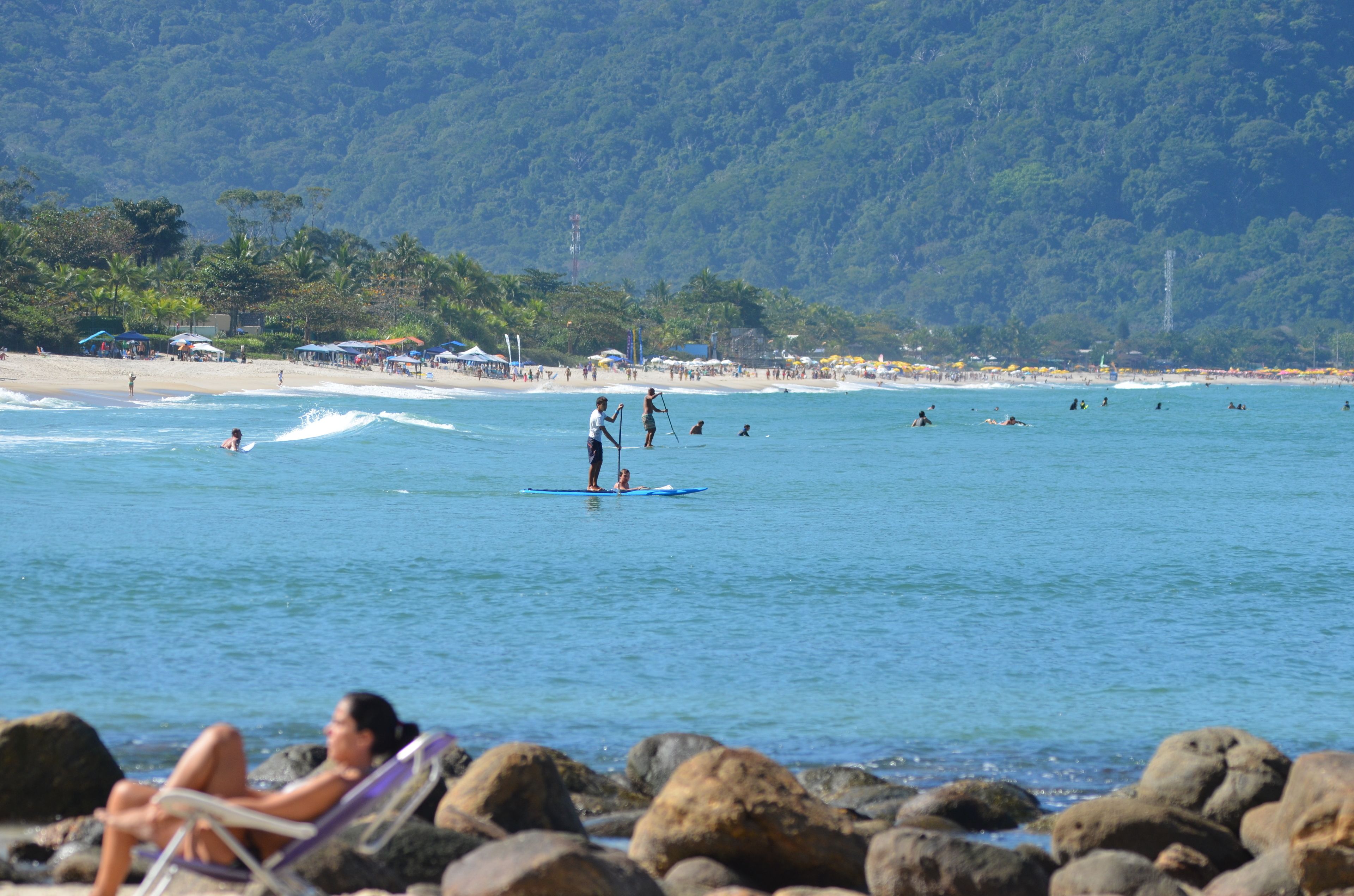 A beach with people engaging in water activities, a rocky shoreline, and a forested hill in the background.