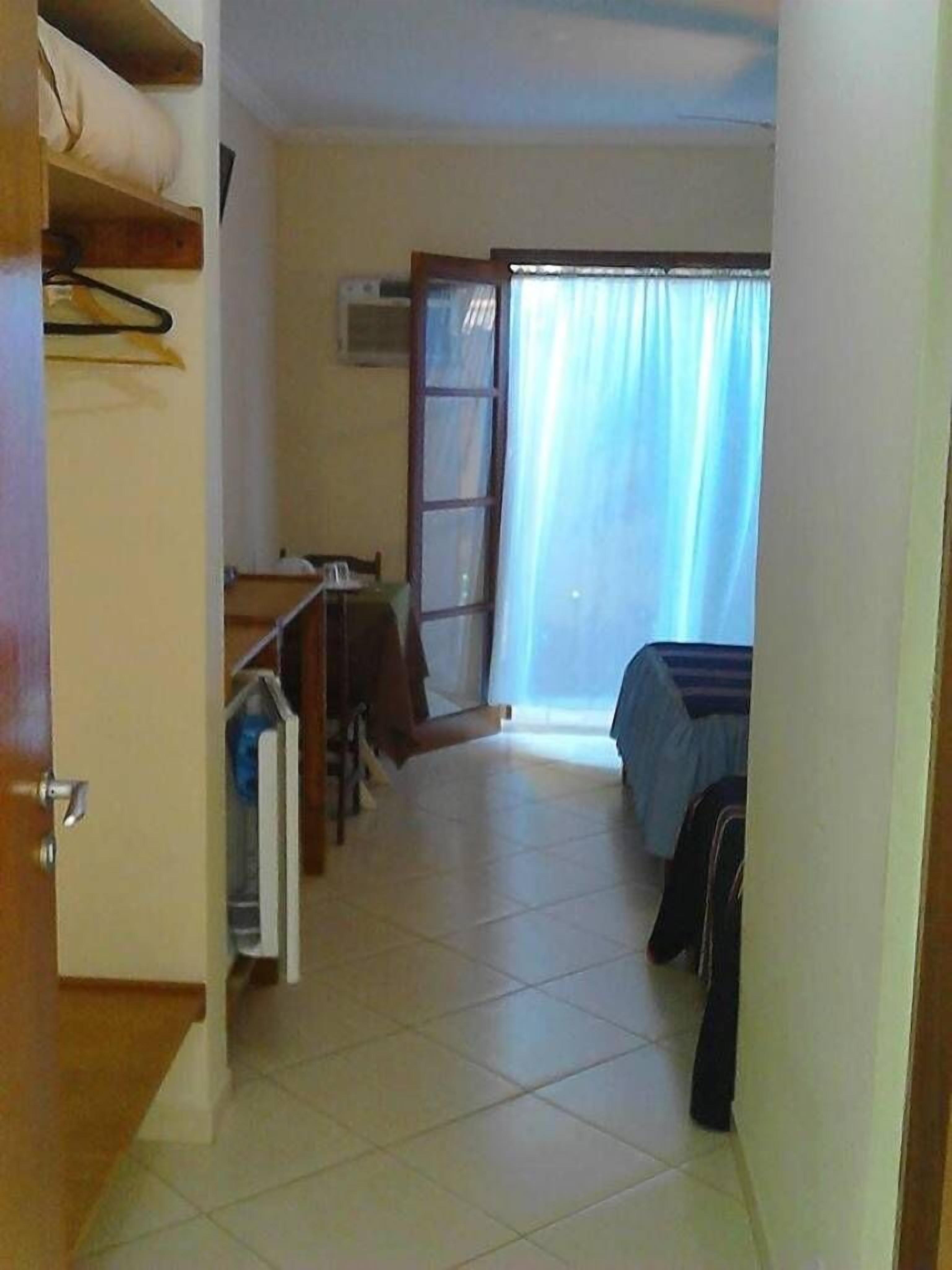 A narrow room with a bed, a desk, and a chair. There is a window with curtains, a door, and a wall-mounted air conditioning unit.