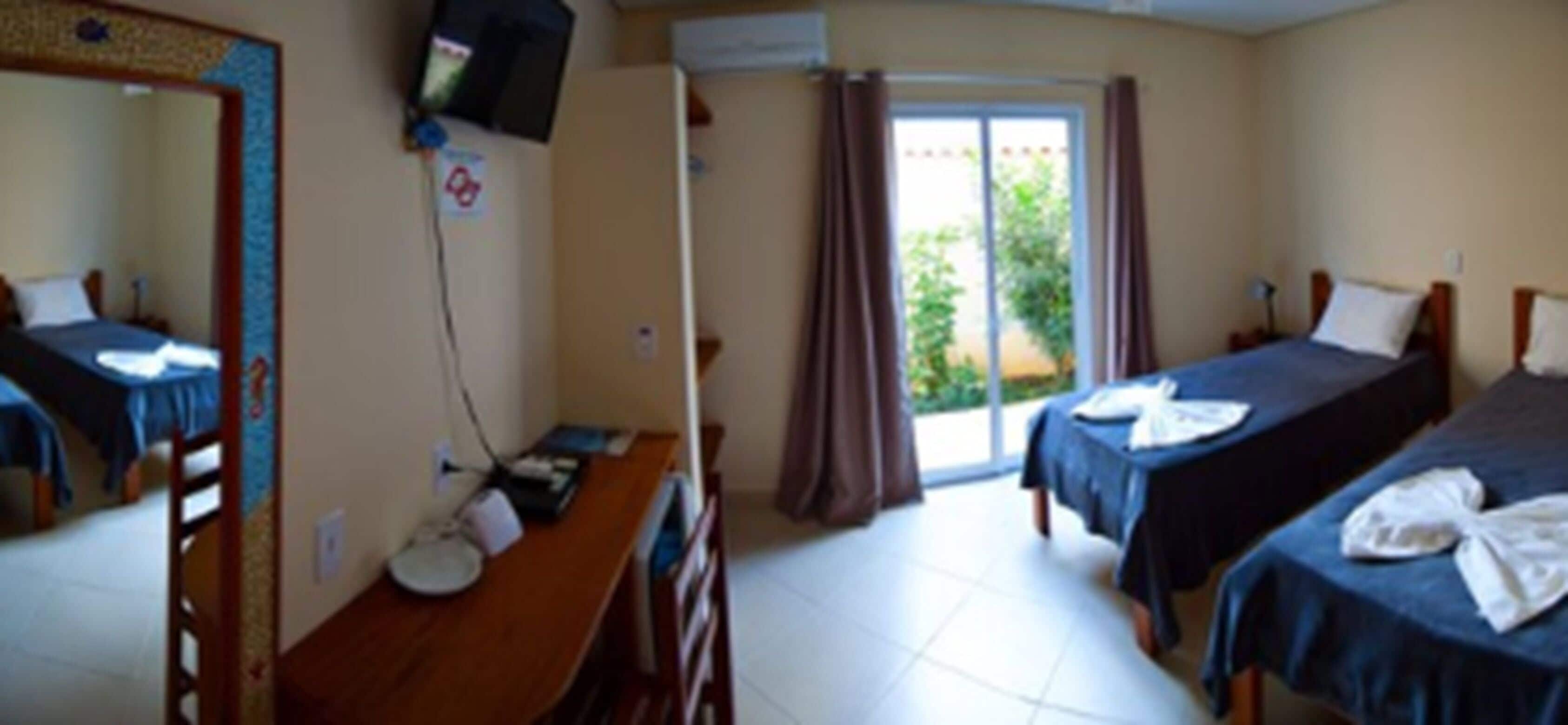 Quarto de hotel com escrivaninha, duas camas, televisão e porta de correr para uma varanda.
