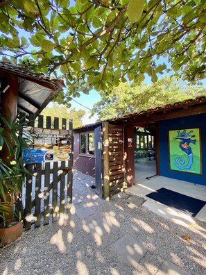 A covered outdoor seating area with a wooden fence, a blue wall with a seahorse mural, and a sign for 'SEU CAFÉ DA MANHÃ'. - Hotel Canto do Rio - Maresias (São Sebastião)