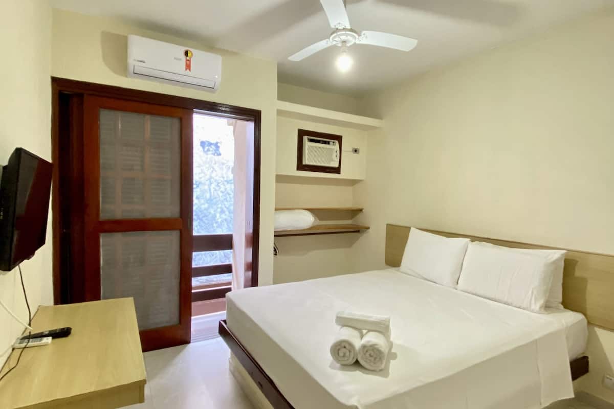 A hotel room with a bed, a ceiling fan, an air conditioning unit, a television, and a wooden desk.