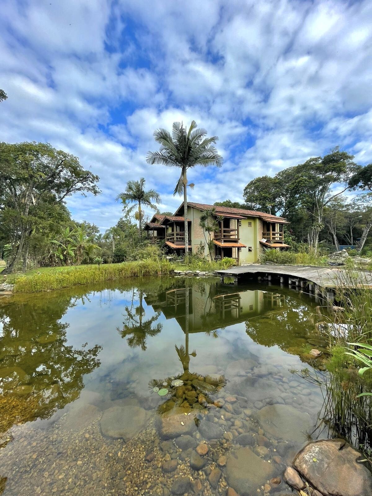 A two-story house with a balcony, surrounded by a pond and lush greenery.