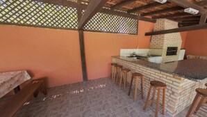 A covered outdoor kitchen area with a brick stove, bar stools, and a counter. - Pousada Mariane (Bombinhas)