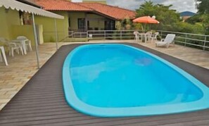 Una piscina rectangular con un borde oscuro, rodeada por una valla blanca y mobiliario de exterior.